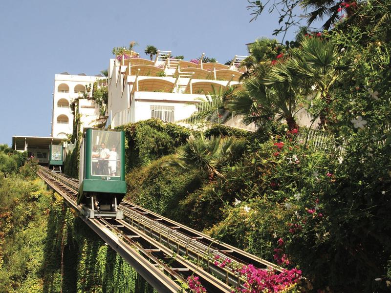 Groene kabelbaanwagen rijdt een spoor omhoog naar een wit hotelgebouw omgeven door groen.