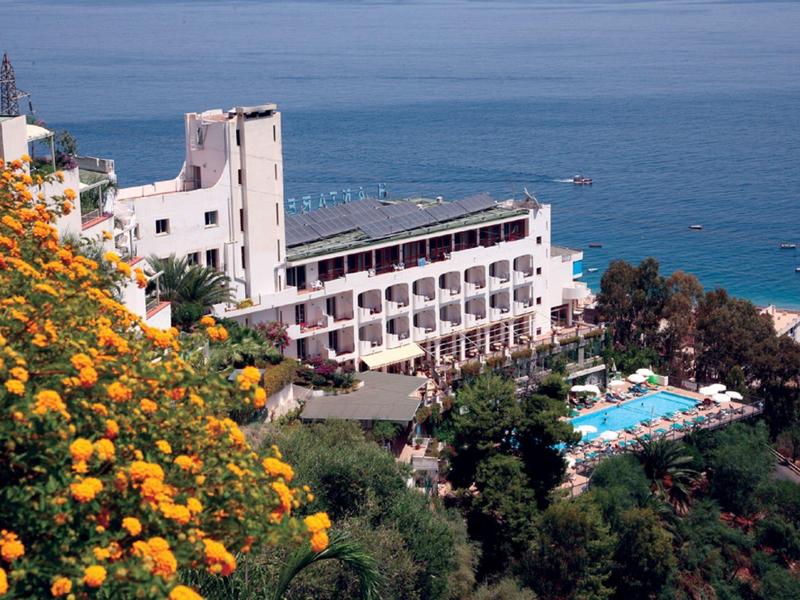 Kusthotel met zwembad, omgeven door groene vegetatie en kleurrijk bloeiende planten.