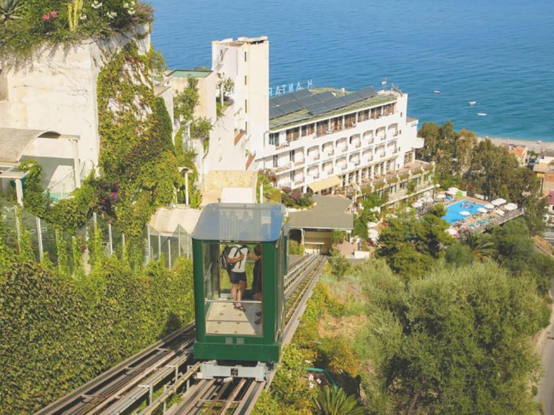 Groene kabelbaan rijdt langs weelderige hellingen met hotel en zee op de achtergrond.