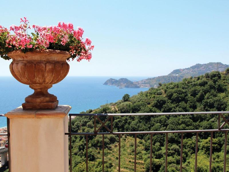 Uitzicht op zee, beboste heuvels en een terras met een bloempot gevuld met roze bloemen.