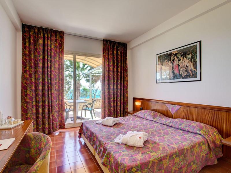 Lichte hotelkamer met tweepersoonsbed, terrasdeur en balkon met tafel en stoelen.