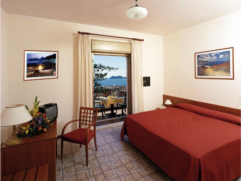 Hotelzimmer mit rotem Bett, Stuhl, Schreibtisch, zwei Bildern und Blick auf Balkon und Meer.