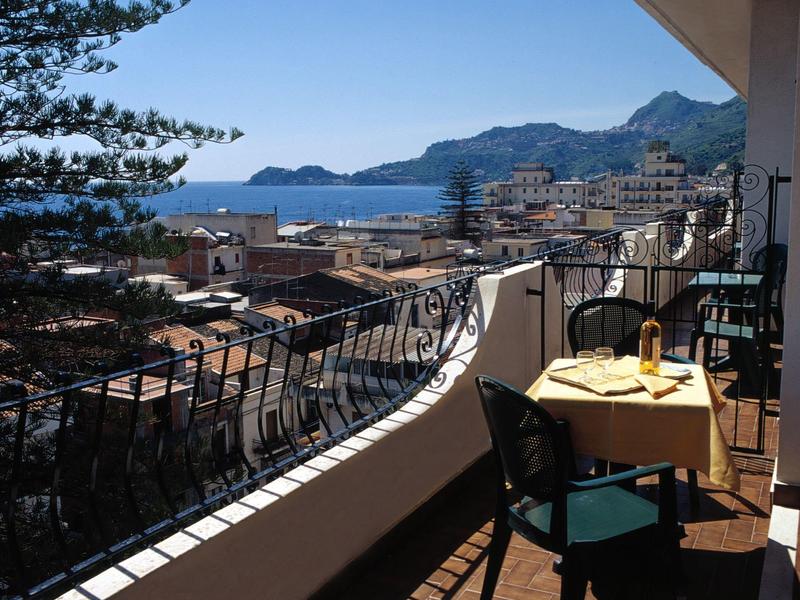Balkon mit Tisch und Stühlen, Blick auf Stadt, Meer und Berge bei klarem Himmel.