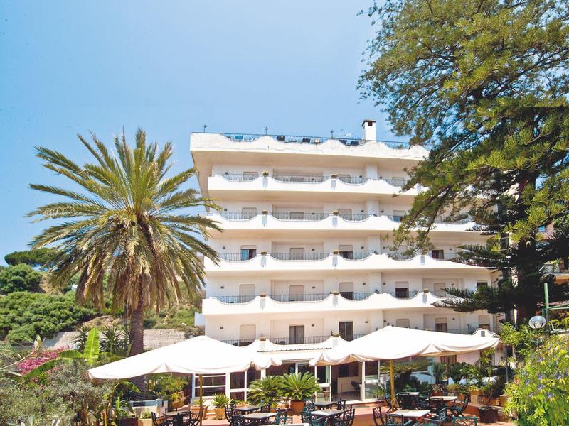 Mehrstöckiges weißes Hotel mit Balkonreihen, umgeben von großen Bäumen und Terrassen mit Tischen und Sonnenschirmen.