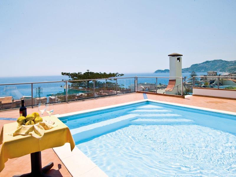 Klare blaue Poolwasserfläche auf Terrasse mit gelb gedecktem Tisch und Meer im Hintergrund.