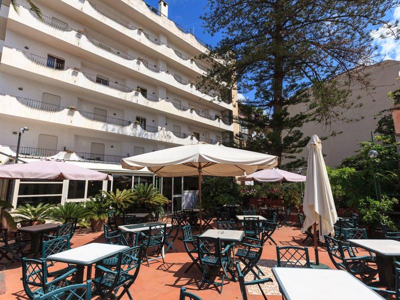 Außenbereich mit Tischen, Stühlen und Sonnenschirmen vor einem mehrstöckigen Hotelgebäude.