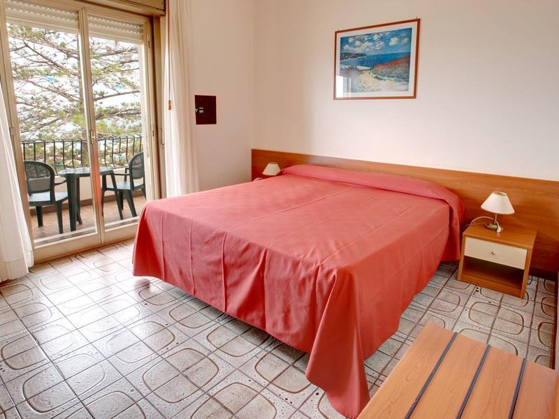 Schlafzimmer mit Doppelbett, roter Bettdecke, Holzmöbeln und Balkon mit Gartenblick.