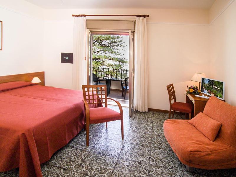 Kleines Zimmer mit Doppelbett, Stuhl, oranger Couch und Balkon mit Blick auf Bäume.