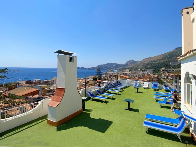 Terrasse mit Liegestühlen, grünem Kunstrasen, Kamin und Blick auf Meer und Berge bei klarem Himmel.