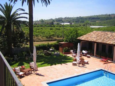 Großer Terrasse mit Sonnenliegen neben Haus, dahinter grüne Wiesen und Palmen unter blauem Himmel.