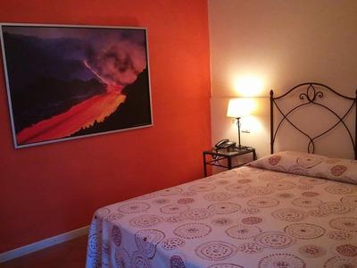 Schlafzimmer mit oranger Wand, gemustertem Bett, Nachttisch, Lampe und Vulkanbild.