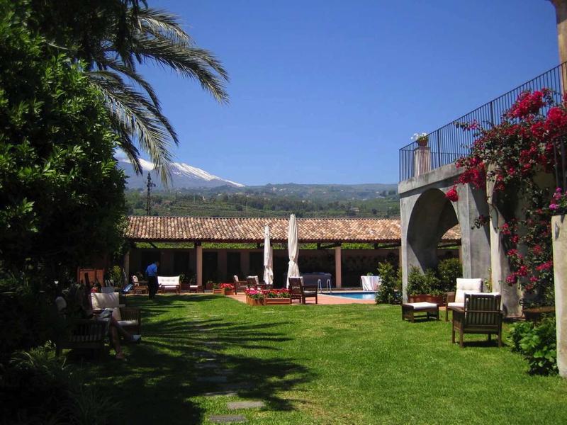 Ein Garten mit grüner Wiese, Stühlen, Palmen, roten Blumen und Berg im Hintergrund unter blauem Himmel.