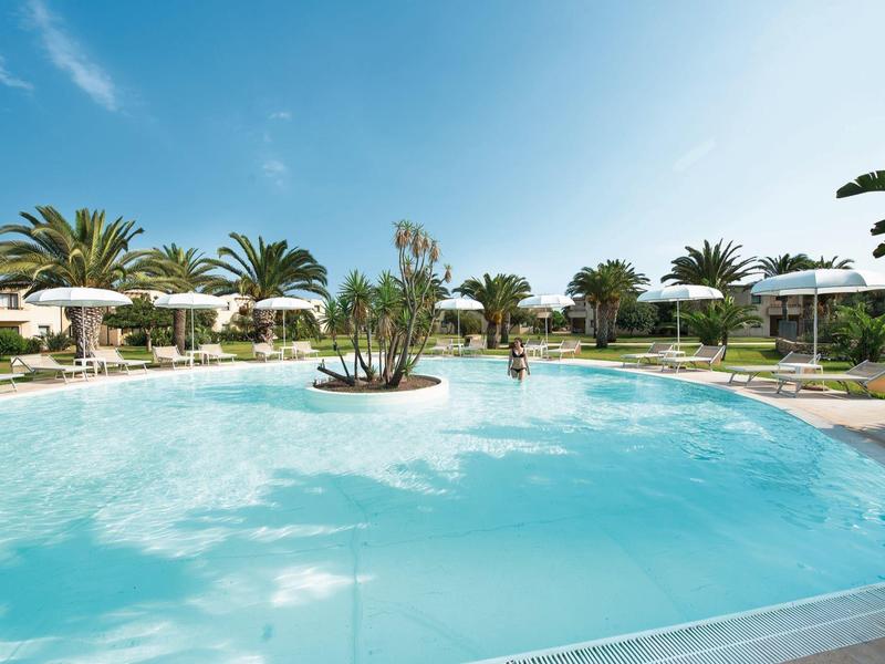 Velký hotelový bazén s palmami, slunečníky a lehátky pod jasnou oblohou.