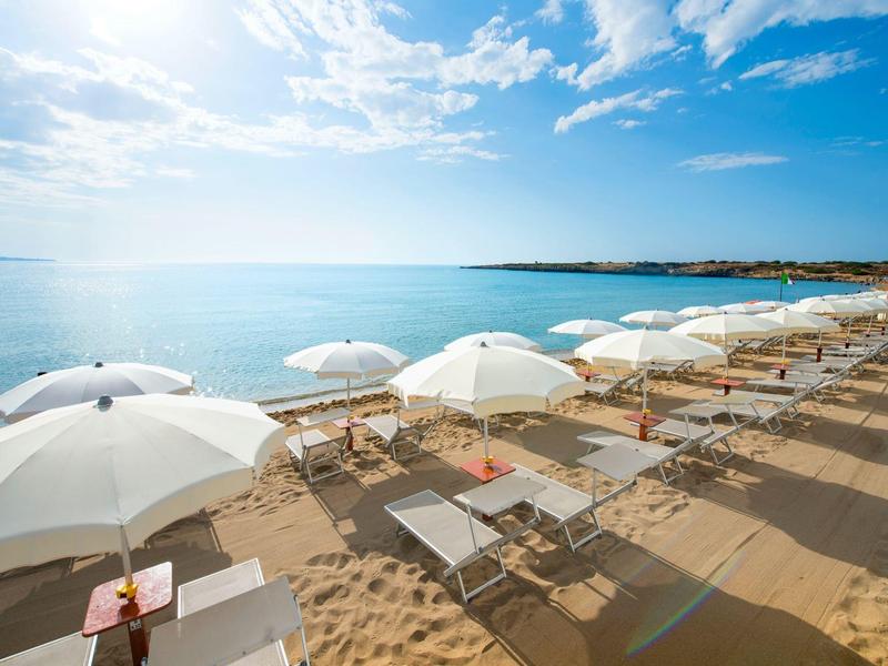 Pláž s bílými slunečníky a lehátky u čistého modrého moře pod slunečnou oblohou.