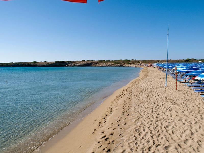 Klidná písečná pláž s průzračnou modrou vodou a řadou modrých slunečníků na pravé straně.