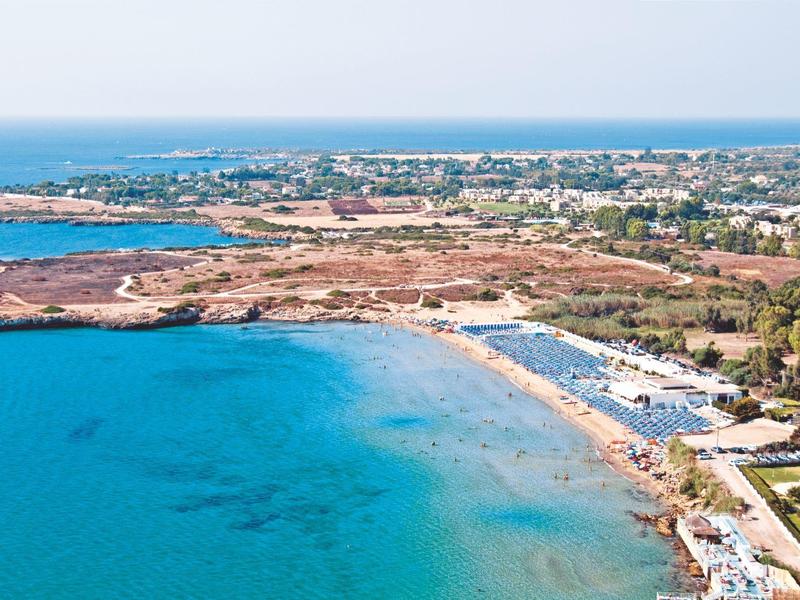 Pobřeží s modrou vodou, pláží a bungalovy pod jasnou oblohou.