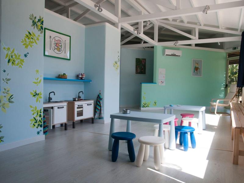 Světla herna se stoly, stoličkami, dětskou kuchyňkou a rostlinnými dekoracemi na stěnách.