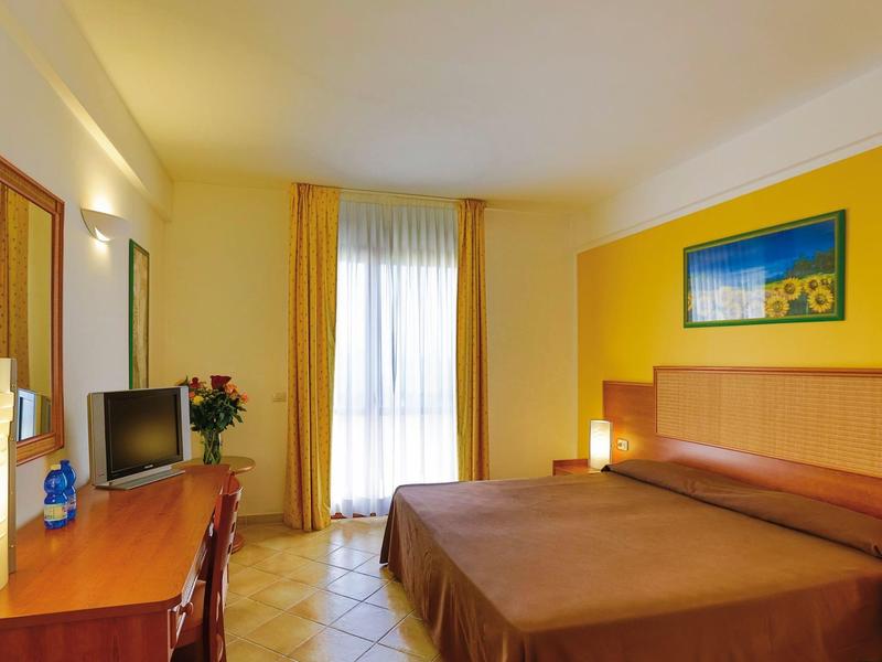 Světly hotelový pokoj s manželskou postelí, psacím stolem, židlí a velkým oknem se žlutými záclonami.