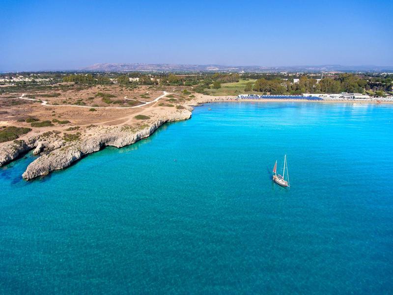 Průzračné tyrkysové moře s plachetnicí a pobřežím pod jasnou oblohou.