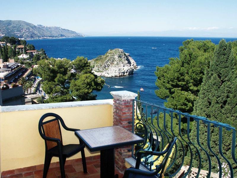 Balkon mit Tisch und zwei Stühlen, grüner Baum und Meer mit Felsen im Hintergrund.