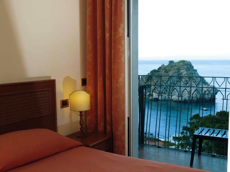 Ein Zimmer mit Bett und Nachttisch, Blick durch offene Tür auf Balkon mit Meer und Felsen.