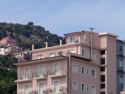 Edificio alberghiero di più piani con balconi davanti a una collina boscosa sotto un cielo azzurro.