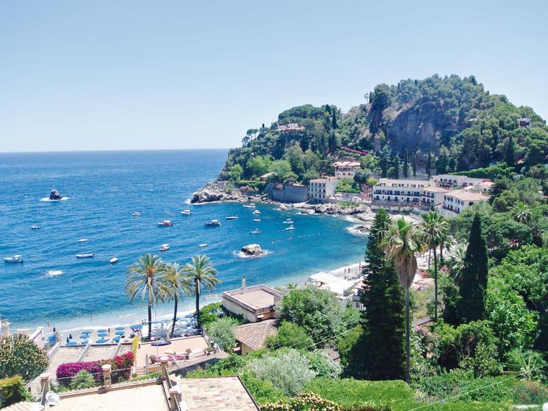 Vista di una costa con una collina boscosa, edifici e barche sul mare blu.