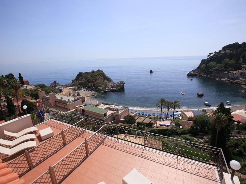 Vista di una cittadina costiera con terrazza, mare e barca sullo sfondo sotto un cielo limpido.