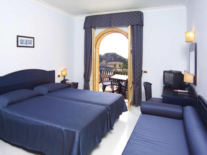 Jednoduše zařízený hotelový pokoj se dvěma jednolůžky, pohovkou a balkonem s výhledem.