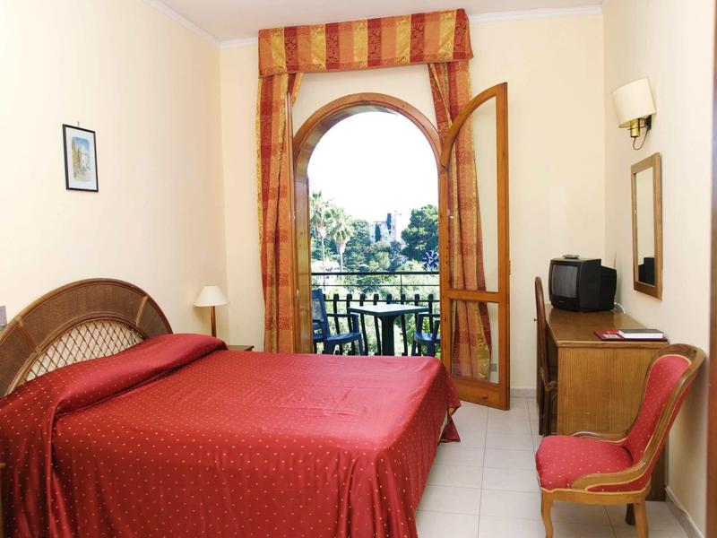 Světle hotelový pokoj s červenou postelí, dřevěným nábytkem a otevřenými obloukovými dveřmi na balkon s výhledem do zeliny.