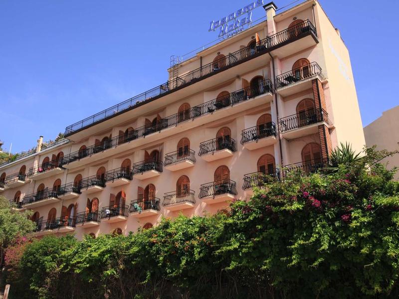 Vícepodlažní budova hotelu s balkony a bujnou zelení pod jasnou modrou oblohou.