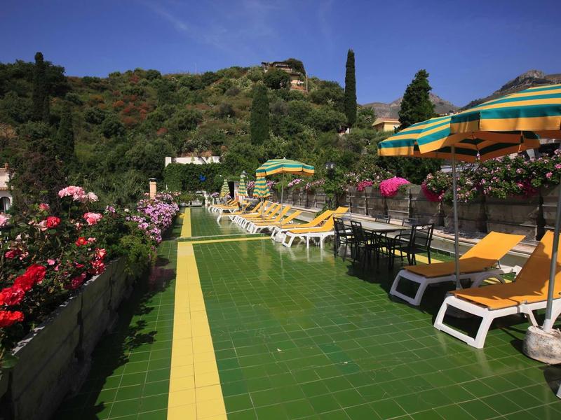 Sluneční terasa se žlutými lehátky, slunečníky a kvetoucími květinami pod jasnou oblohou.