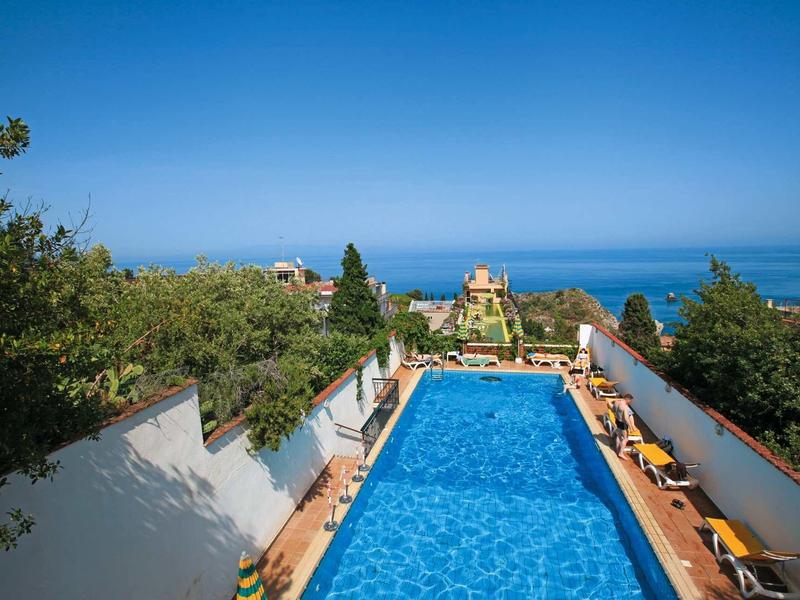 Pohled na modrý bazén s lehátky a moře v pozadí.