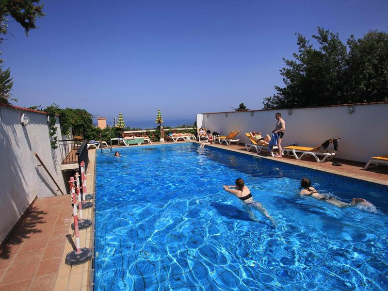 Pravoúhlý venkovní bazén s lehátky a lidmi, modrá obloha a moře v pozadí.