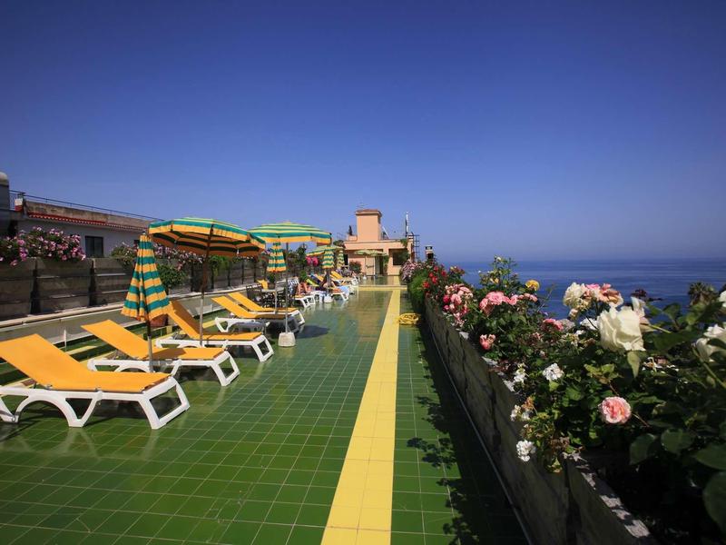 Střešní terasa s lehátky a květinami, výhled na moře za jasné oblohy.