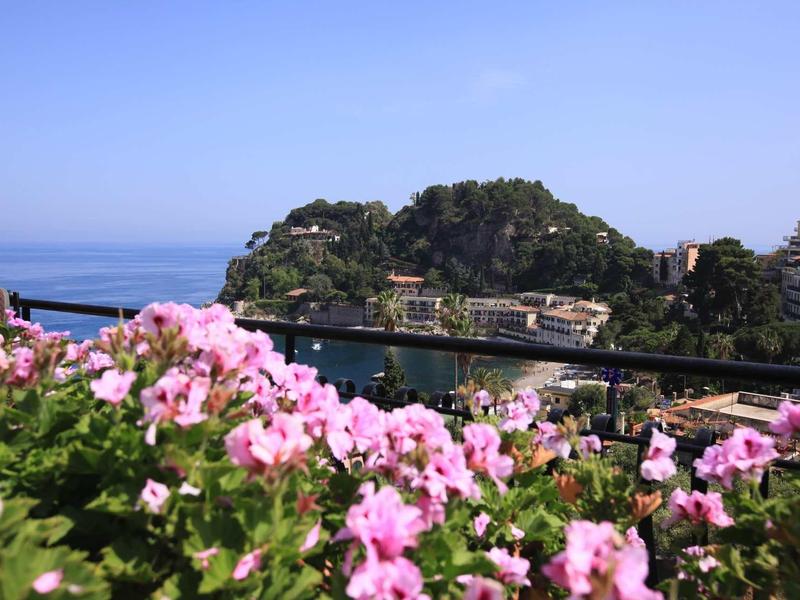 Pohled na zátoku s růžovými květy v popředí a kopci v pozadí pod jasnou oblohou.