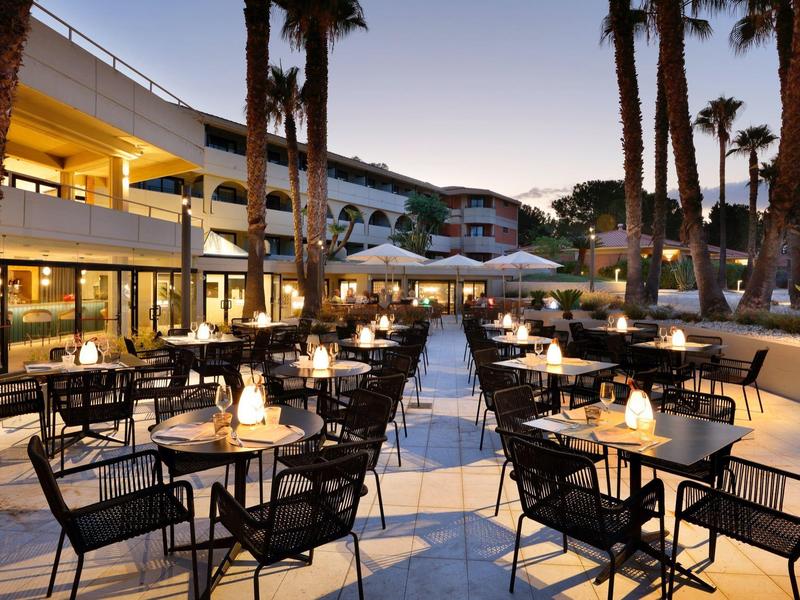 Outdoor hotel restaurant seating with tables, chairs, palm trees, and evening lights.