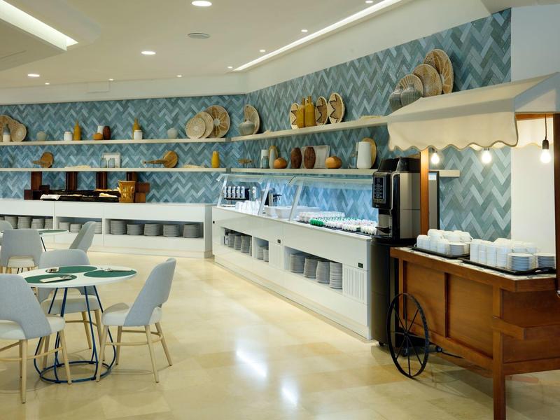 Bright hotel breakfast area with buffet counters, shelves of plates, and modern seating.