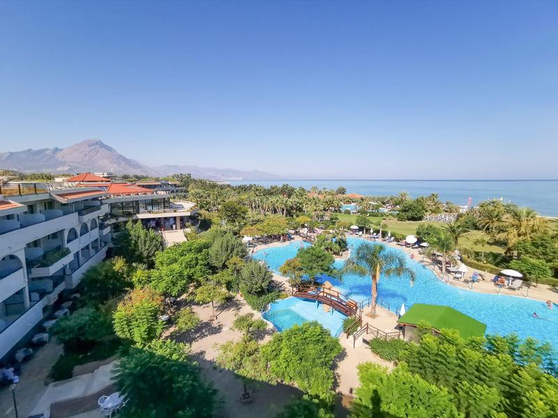 Resort with large pool, surrounded by greenery and river sea in the background under clear sky.