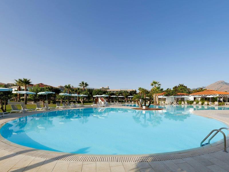 Large outdoor pool with clear water surrounded by sun loungers and umbrellas under a blue sky.
