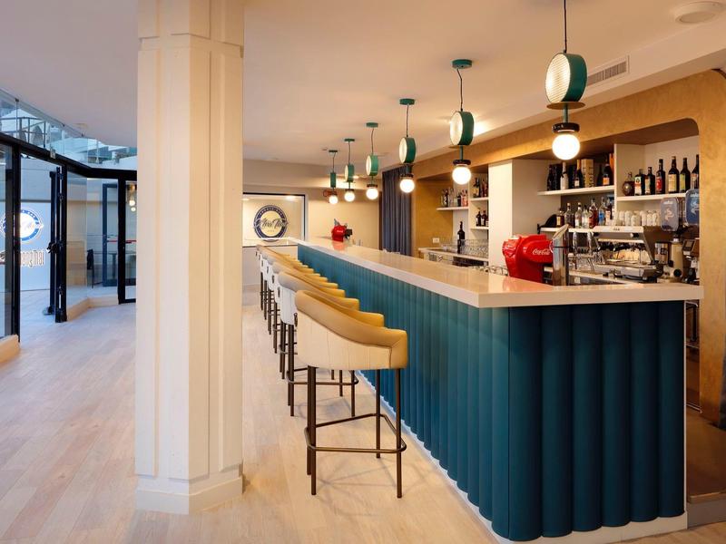 Modern hotel bar with blue counter, high stools, and pendant lights during daytime.