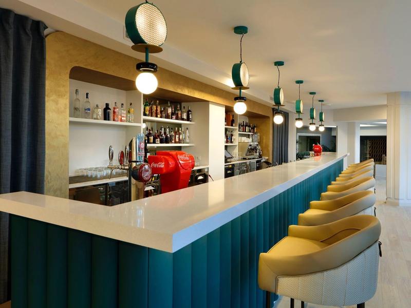 Modern hotel bar with a long counter, yellow chairs, hanging lights, and shelves with drinks.