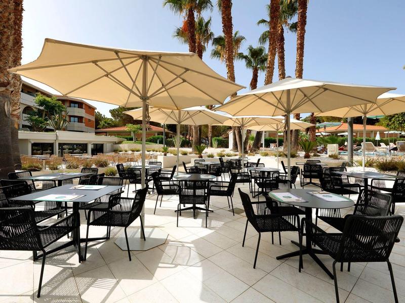 Outdoor hotel patio with black chairs and beige umbrellas under palm trees on sunny day.