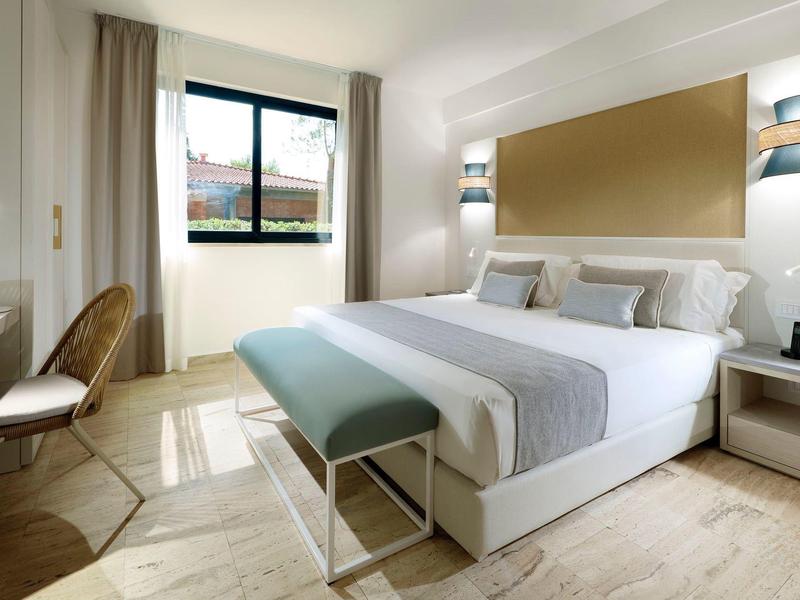 Bright hotel room with queen bed, blue bench, wooden chair, and large window.