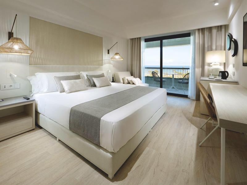 Modern hotel room with king bed, ocean view, light wood flooring, and minimalist decor.