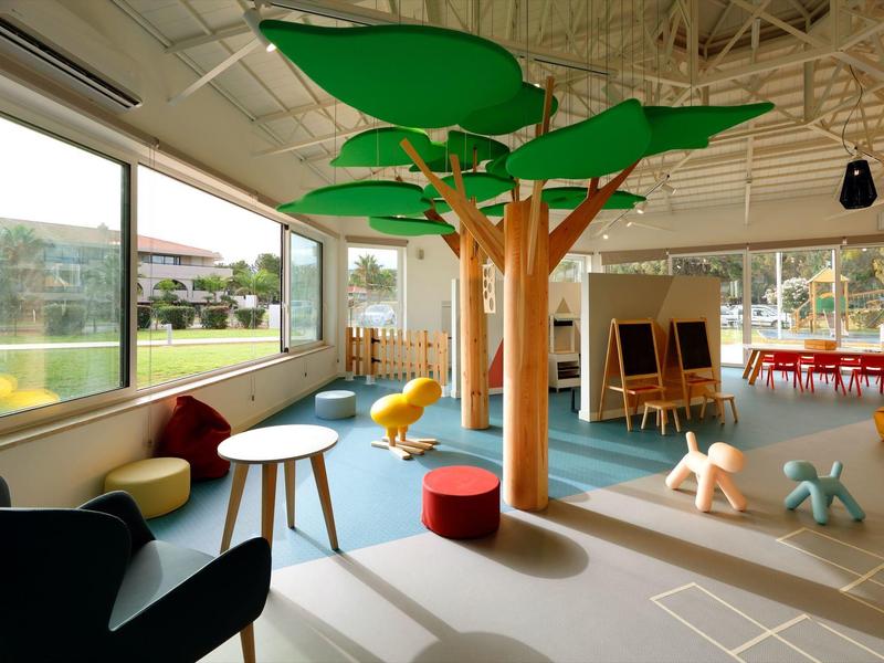 Bright indoor play area with artificial trees, colorful animal seats, and large windows overlooking greenery.