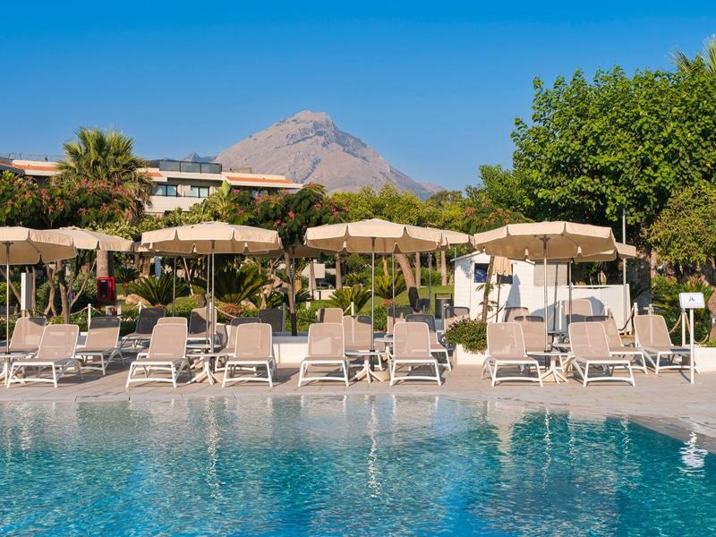 Poolside lounge chairs under umbrellas with a mountain in the background on a sunny day.