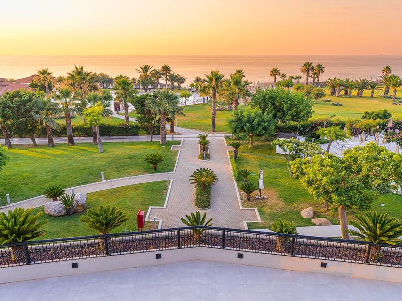 Sunset view of a lush garden with palm trees and walking paths by the sea.