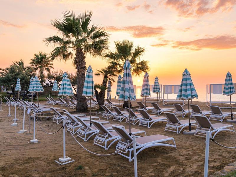 Empty beach chairs with striped umbrellas at sunset near palm trees and calm sea.
