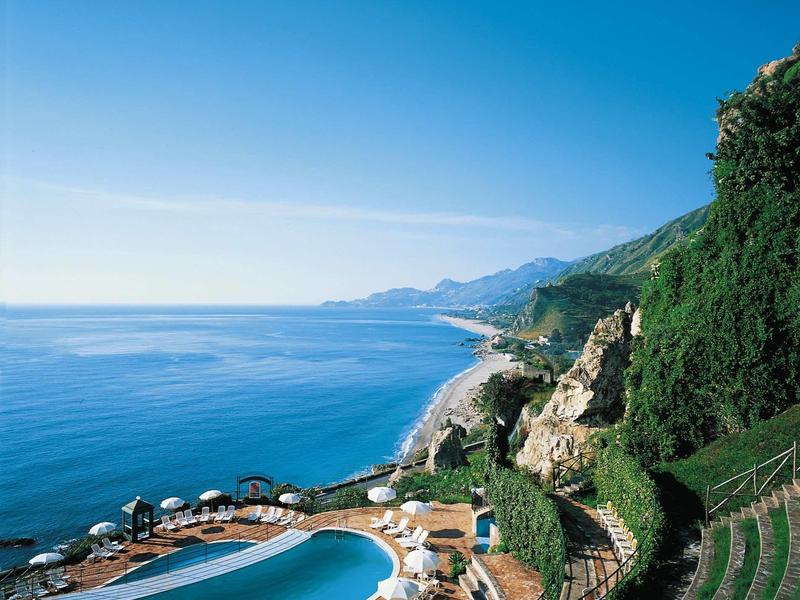 Zwembad met ligstoelen aan de kust met uitzicht op de blauwe zee en groene heuvels.